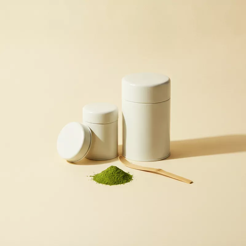Eine kleine und eine große cremeweiße Teedose, davor Matcha-Pulver und ein Bambuslöffel