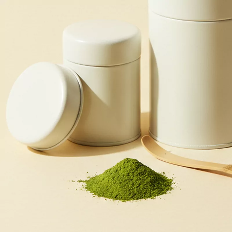 Kleine cremeweiße Teedose mit abgenommenem Deckel, davor ein Häufchen leuchtend grünes Matcha-Pulver