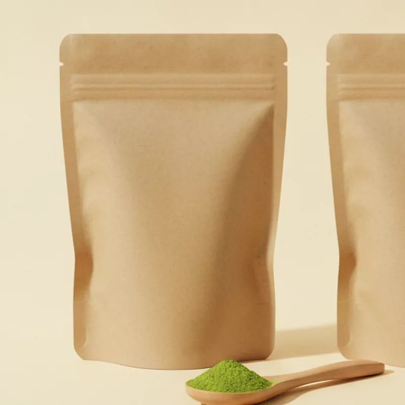Standbeutel aus Kraftpapier, davor ein Holzlöffel mit grünem Matcha-Pulver