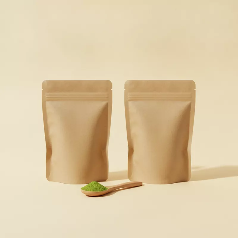 Zwei Standbeutel aus Kraftpapier nebeneinander, davor ein Holzlöffel mit Matcha-Pulver