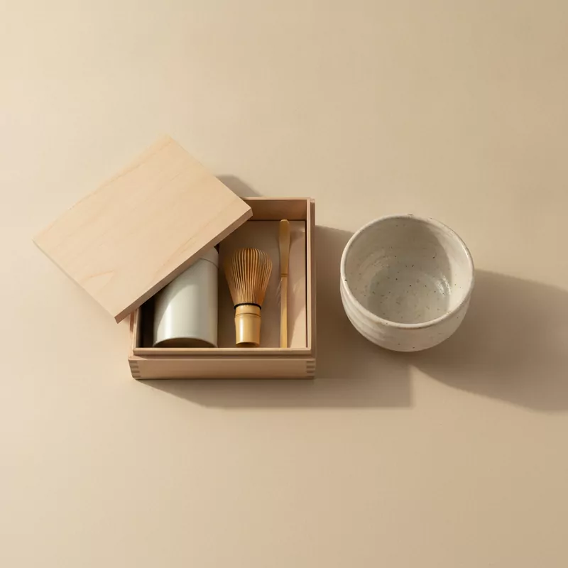 Offene Holzschachtel mit Teedose, Bambusbesen und Bambuslöffel, daneben eine cremeweiße Matcha-Schale
