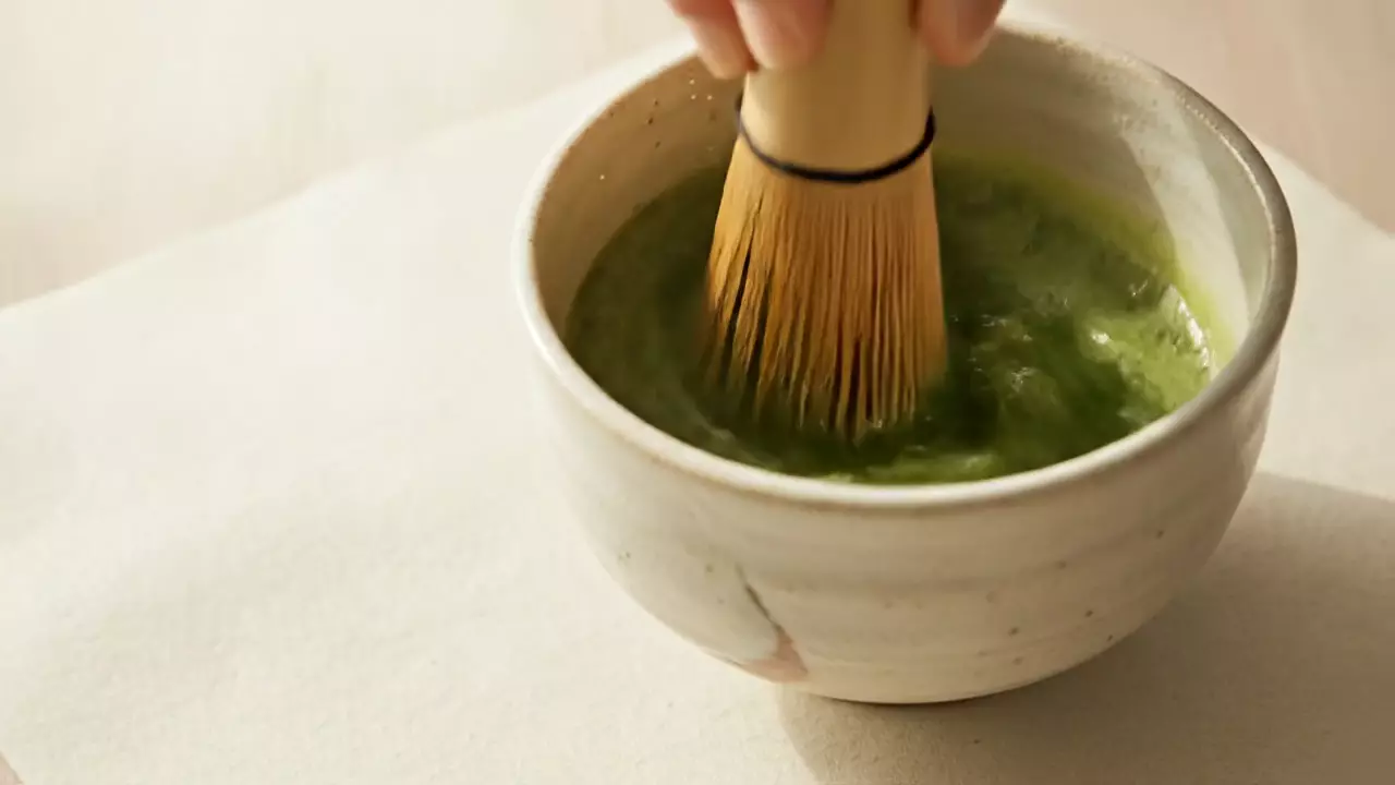 Ein Bambusbesen schlägt Matcha in einer cremeweißen Keramikschale auf, auf der Oberfläche bildet sich feiner hellgrüner Schaum